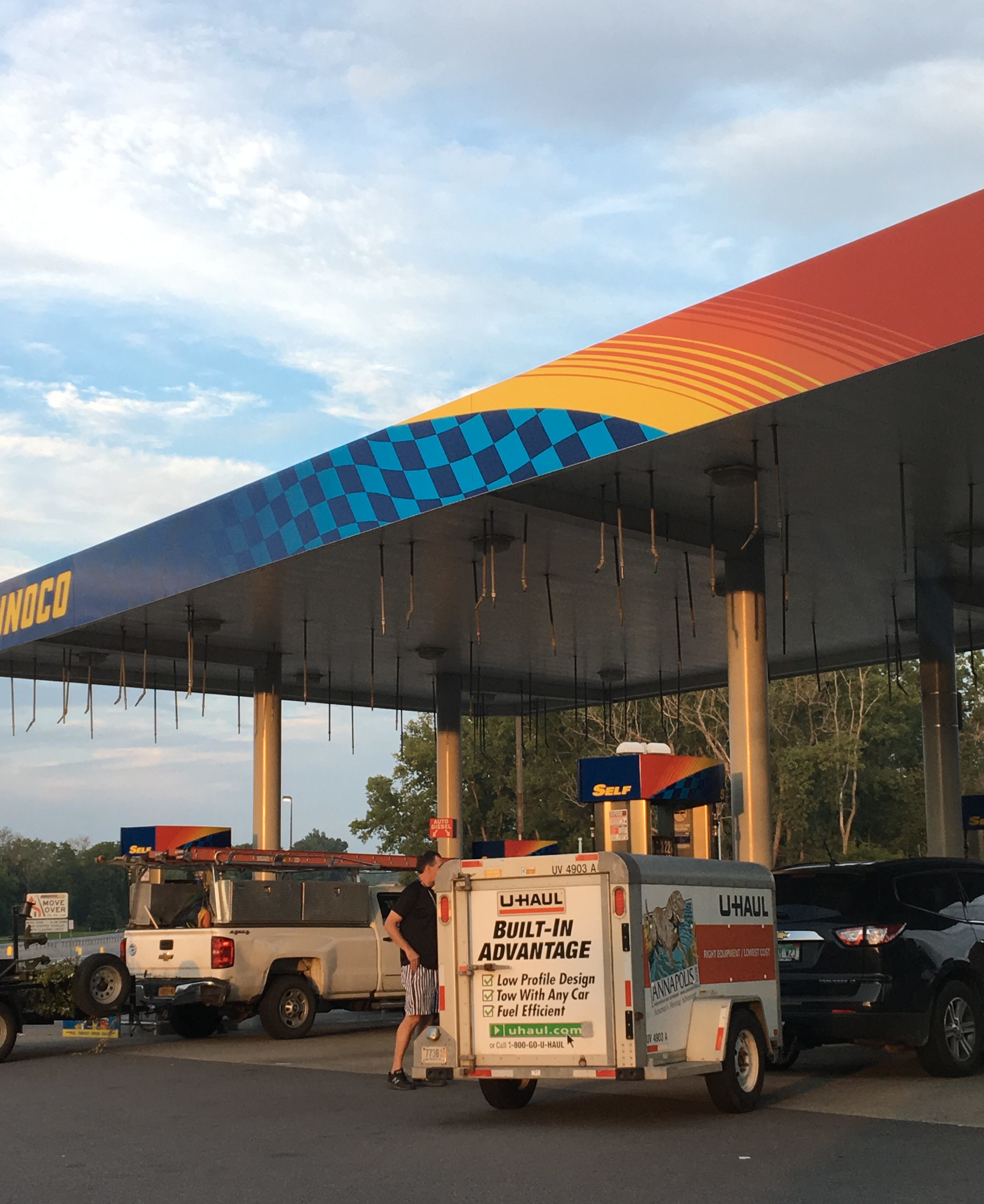 An image of a gas station. In front of it, attached to a truck which is out of frame, is a U-HAUL box smaller than the average car.