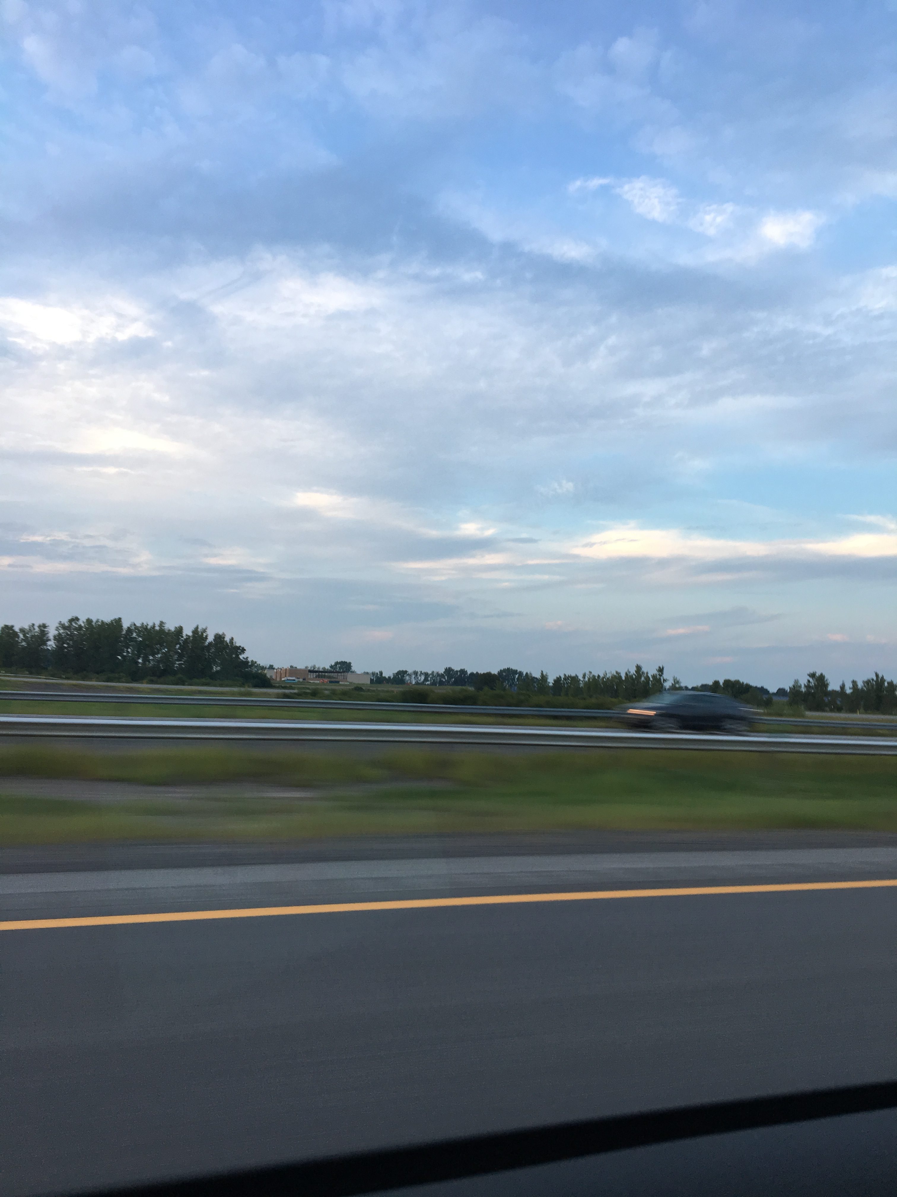 A view of the sky, with blurry trees and road at the bottom of the photo. The sky is light blue with lots of clouds in various shades of white.