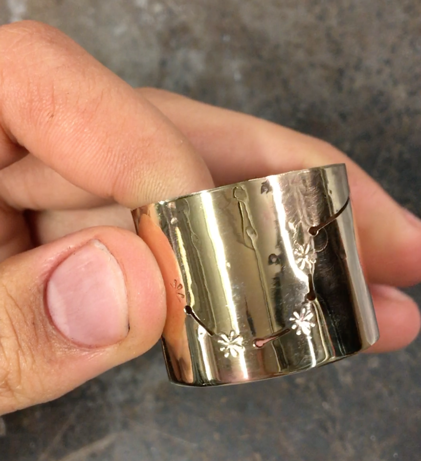 My hand holding a small cylinder of polished, shiny metal. It has imprinted asterisks connected by cut-out lines, meant to resemble constellations.