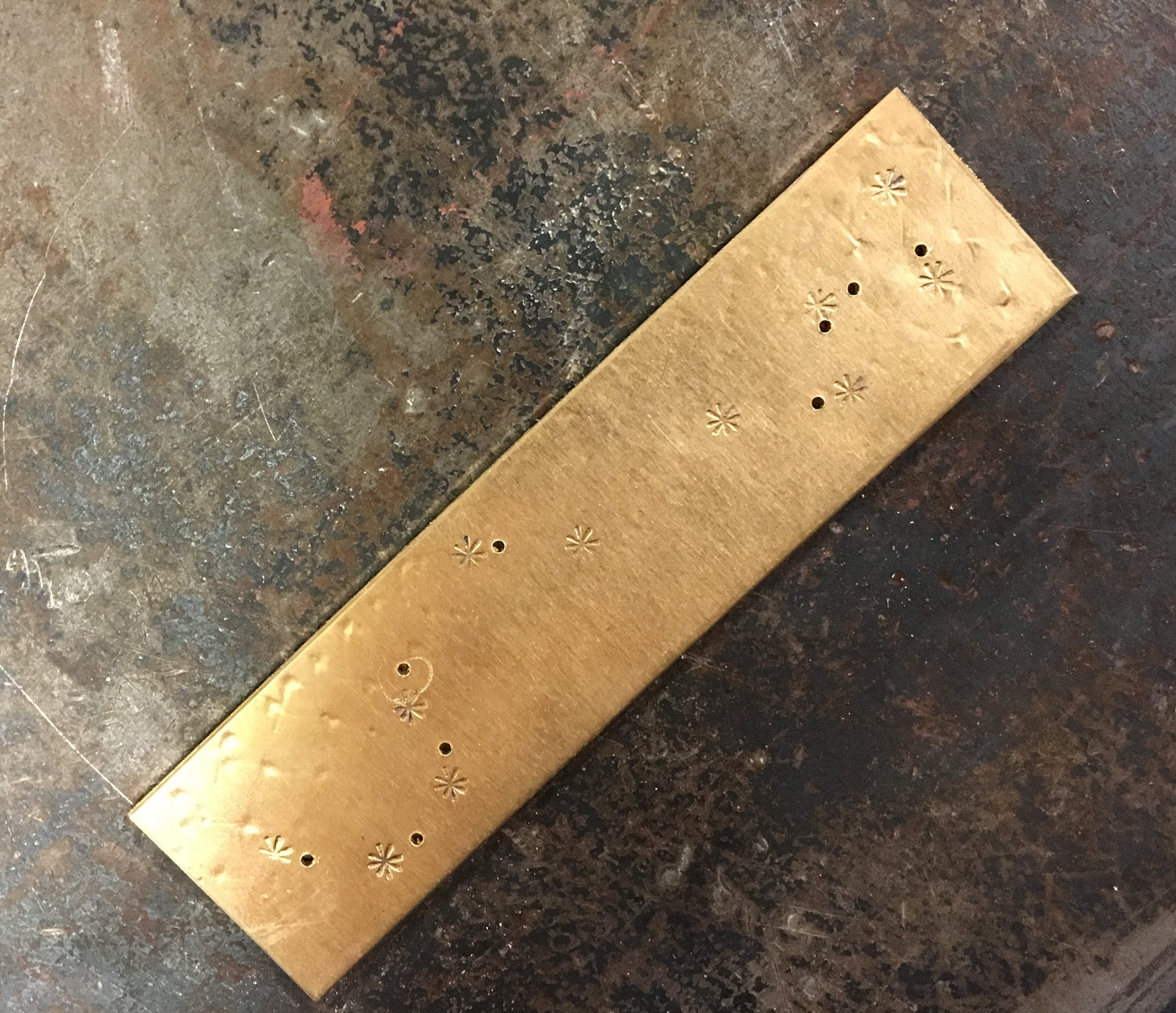 A flat rectangle of yellow, slightly shiny copper. At the top edge faint indentations can be seen; patterned along the middle are asterisks and small holes next to them.