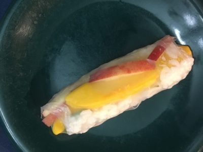 A finished summer roll on a small plate. The mango, apple, and rice are visible through the clear rice paper.