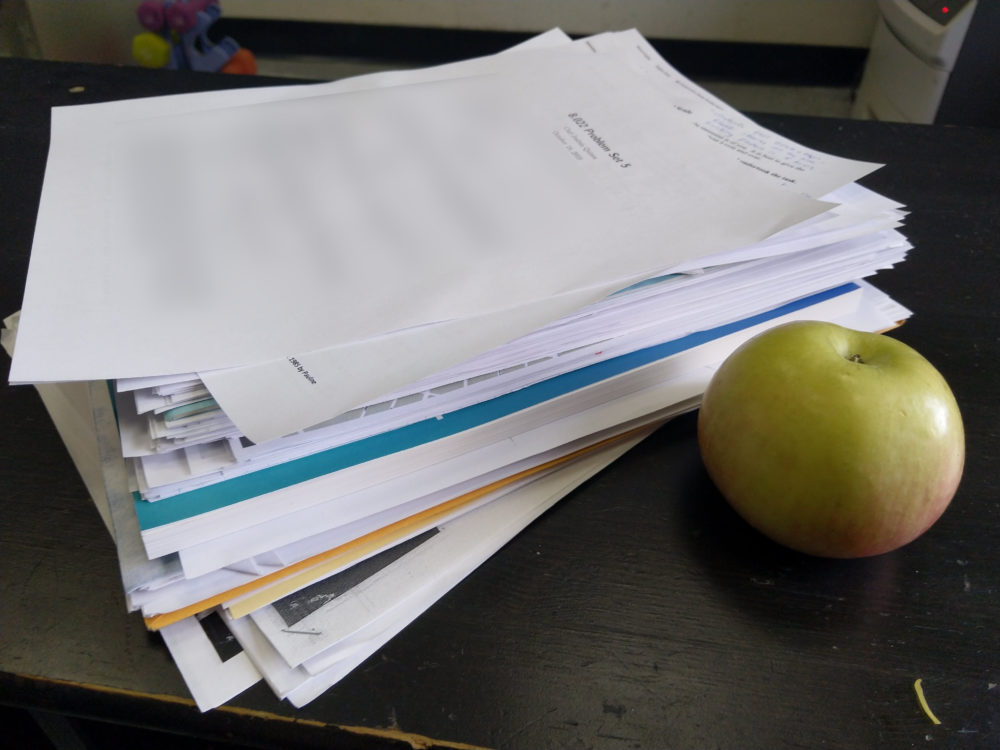 a stack of paper. next to it is an apple. the stack of paper is about twice as tall as the apple.