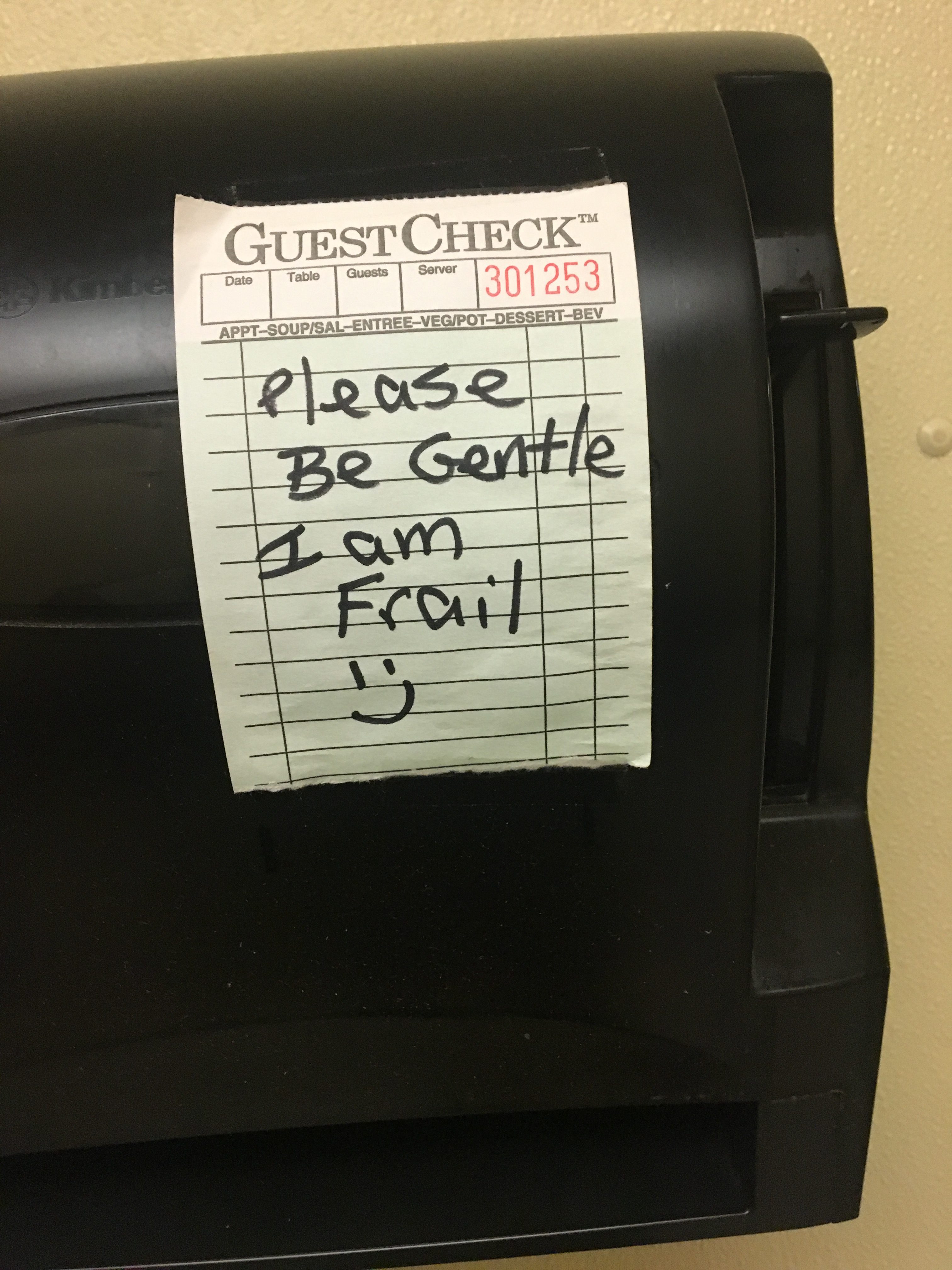 A paper towel dispenser in a restaurant. A small sign on it reads "Please Be Gentle I am Frail =)".