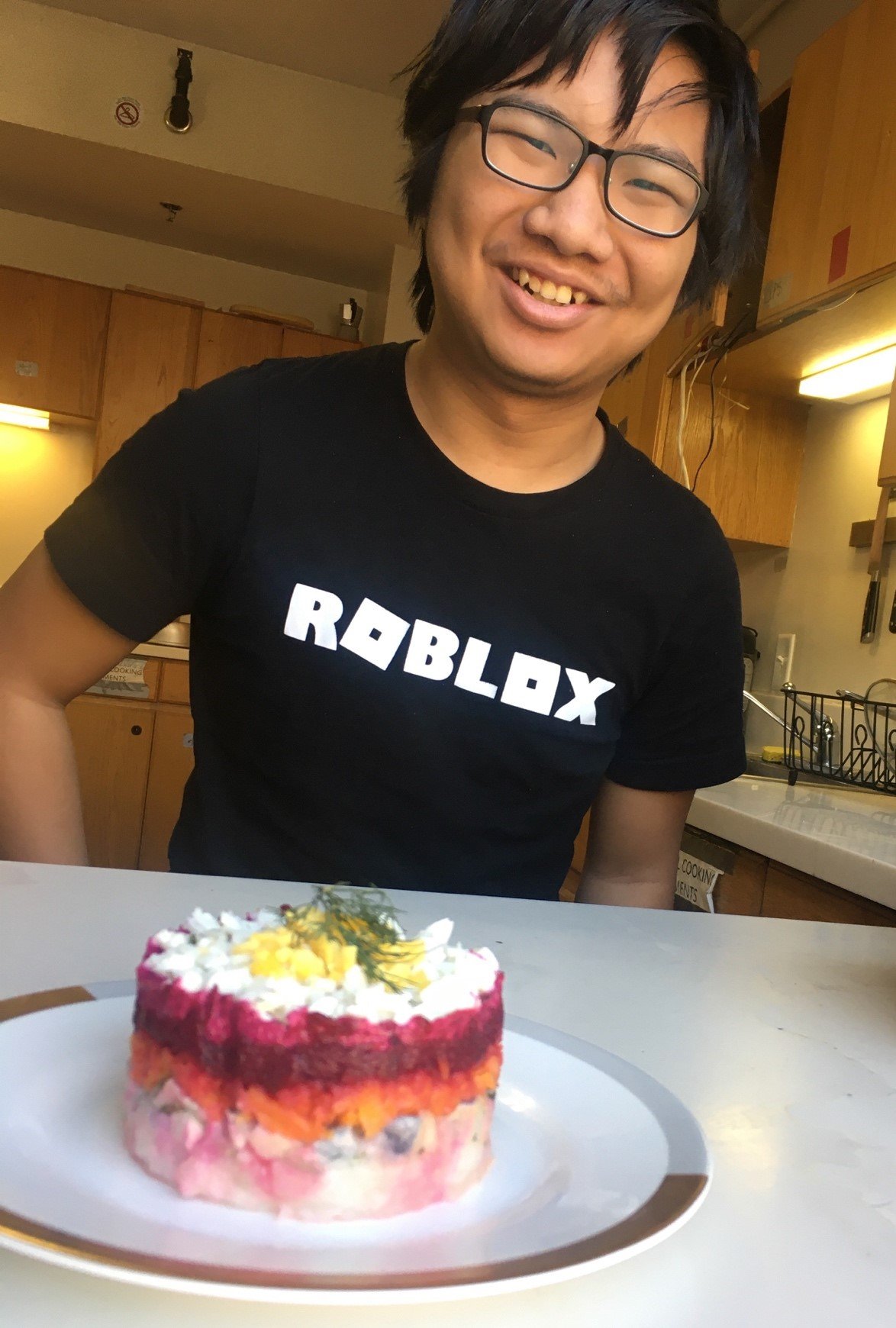 me, with a roblox shirt, in background. in foreground is a plate, with a dish on it, made of several layers of vegetables and fish.