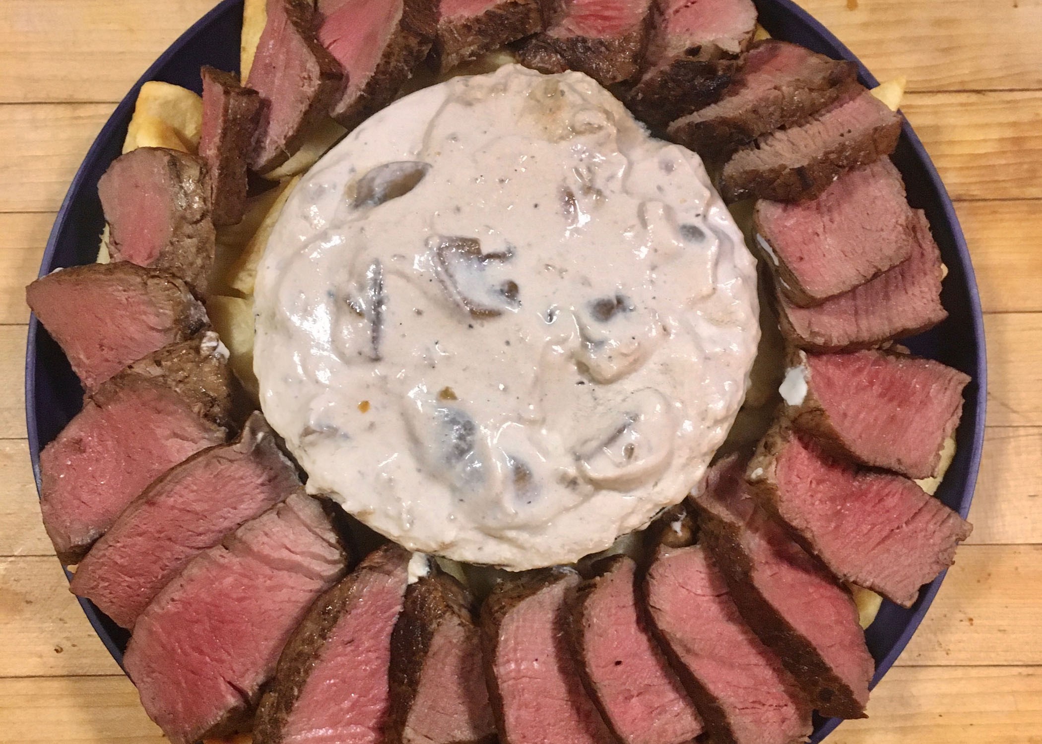 a plate of sliced beef, on top of potatoes, with a bowl of mushroom sauce in the middle