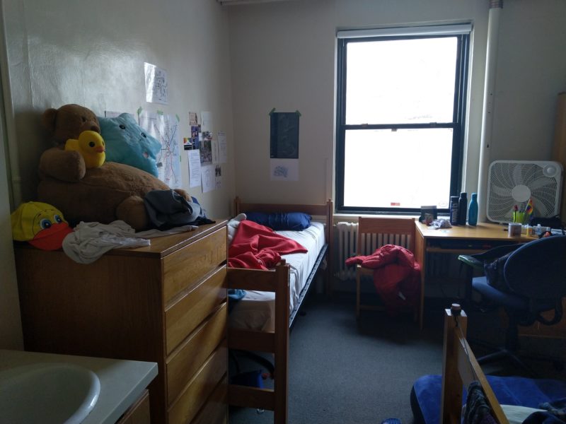my half of the room. a cabinet in the foreground with stuffed animals on top of it, a bed behind. visible posters on top of the bed. a desk underneath a window.