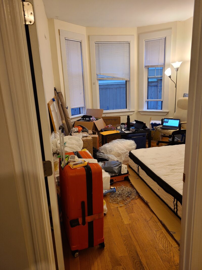 queen mattress on ground with very messy boxes and suitcases