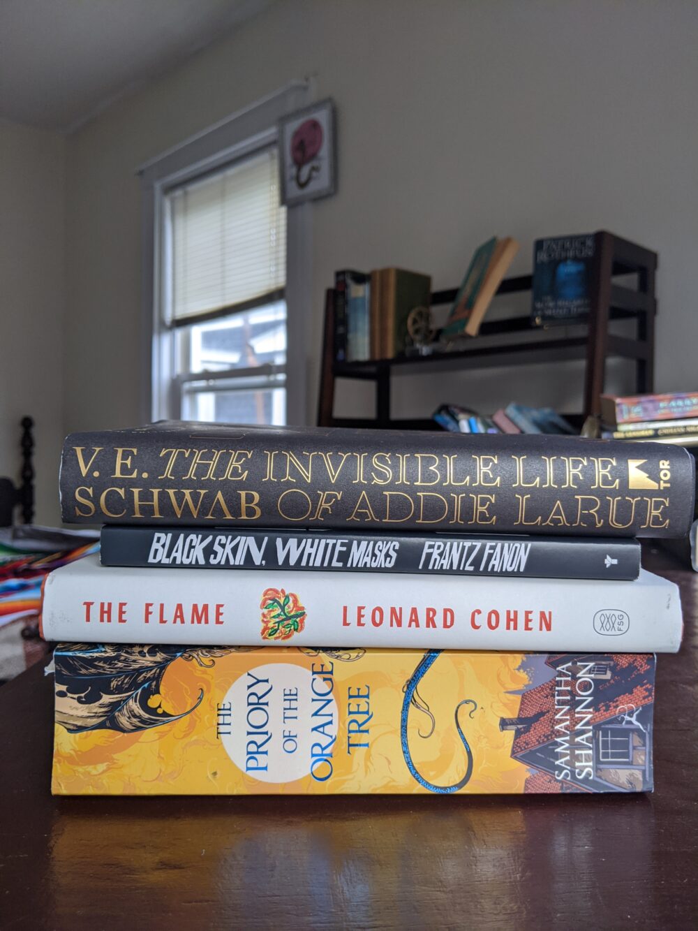 stack of books: The Invisible Life of Addie LaRue by VE Schwab, Black Skin, White Masks by Franz Fanon, The Flame by Leonard Cohen, and Priory of the Orange Tree by Samantha Shannon