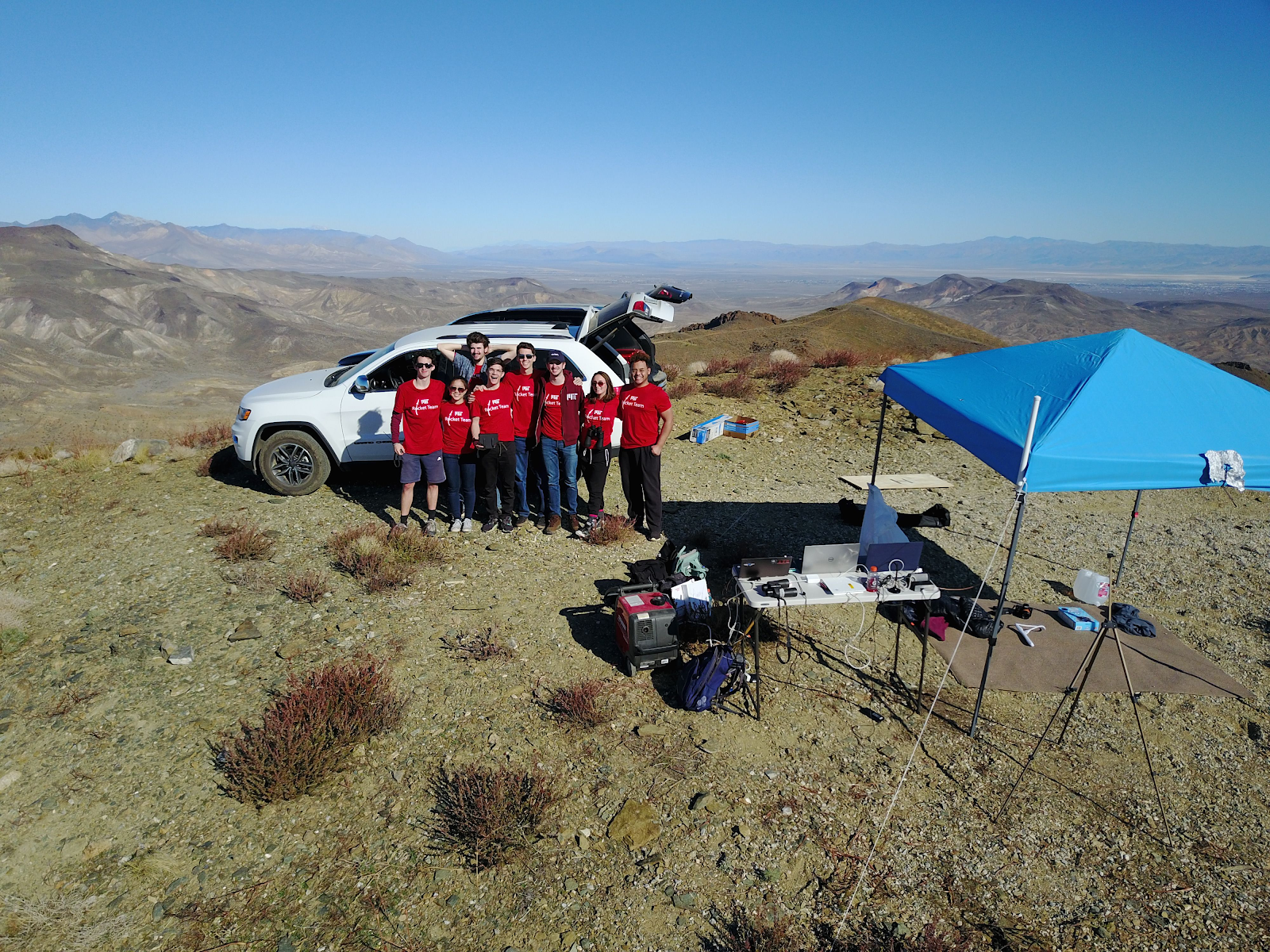 another group picture of the rest of Rocket Team standing in front of a car near an Avionics table and tent on a mountaintop in the Mojave Desert