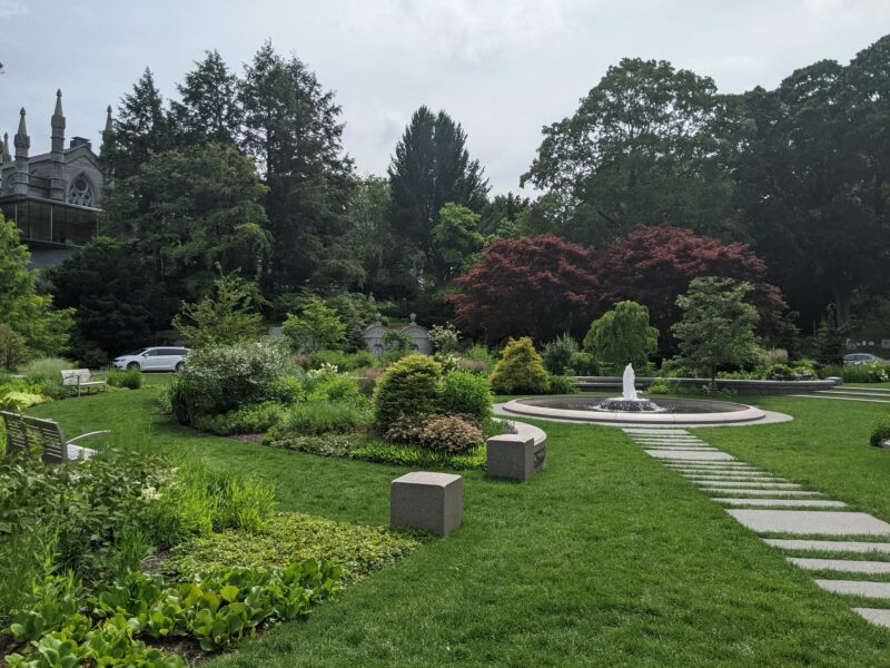 small fountain surrounded by bushes, with trees and a chapel behind them