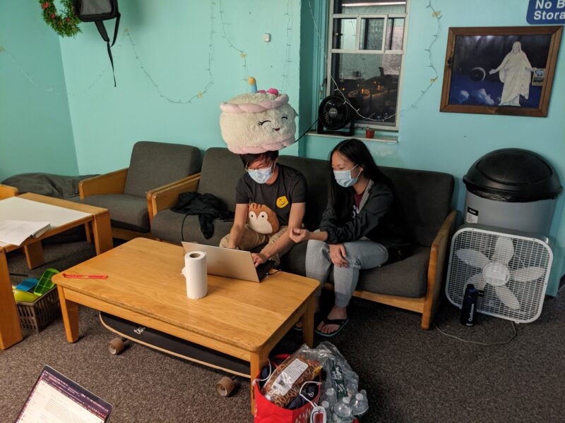 two people sitting on a couch; one has a large cake plushie on their head