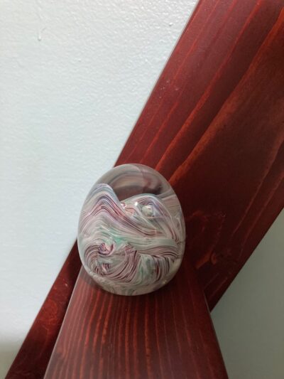 An egg-shaped glass paperweight. It is sitting on a maroon ladder in front of a green wall. The paperweight itself has rolling hills inside it made up of streaks of maroon, green, and white color.