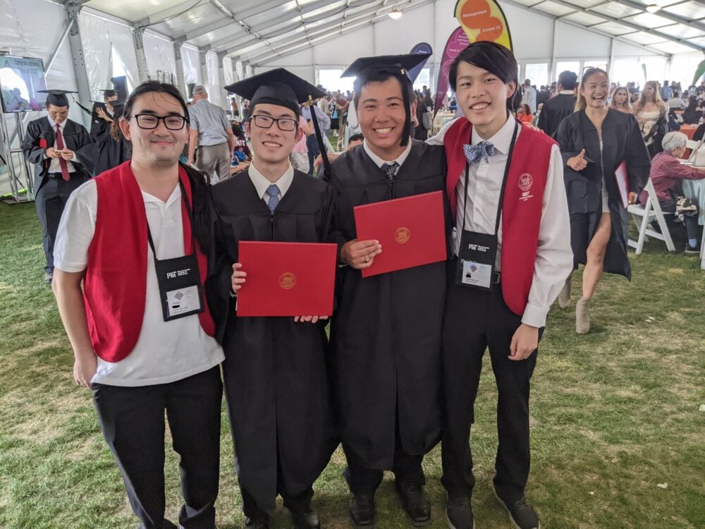 four individuals: two in graduation robes, two in volunteer vests, smiling for a picture