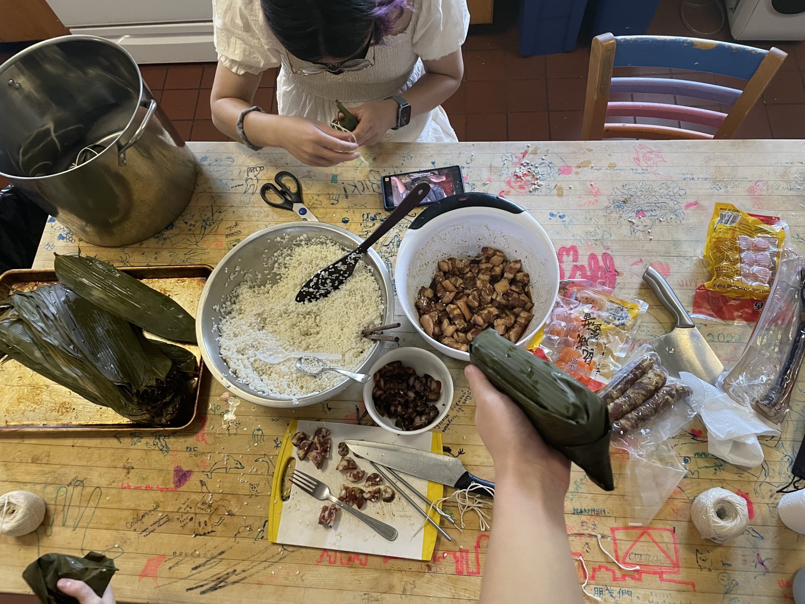 Winnie wraps a zongzi next to a pot while watching a tutorial online. Alice is holding a somewhat ugly zongzi in the foreground.