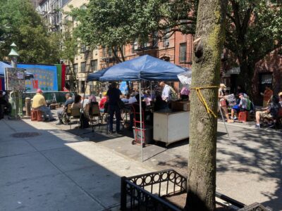 A small street. There is a stage set up in the street with no one on it. A couple dozen people sit on folding chairs in front of the stage.