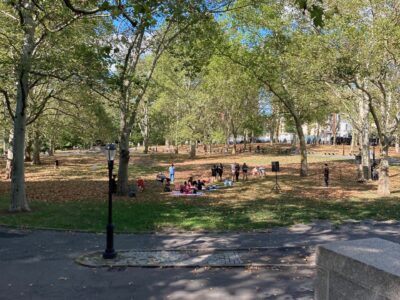 A park with grass and fallen leaves. There are a few small groups of people sitting on the grass and one slightly larger group with a speaker next to them.
