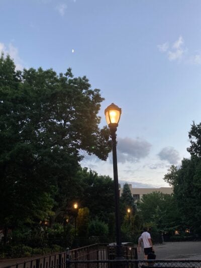 A park at dusk. The sky is light blue, but it's dark enough that the streetlight in the middle of the photo is lit up and glowing.