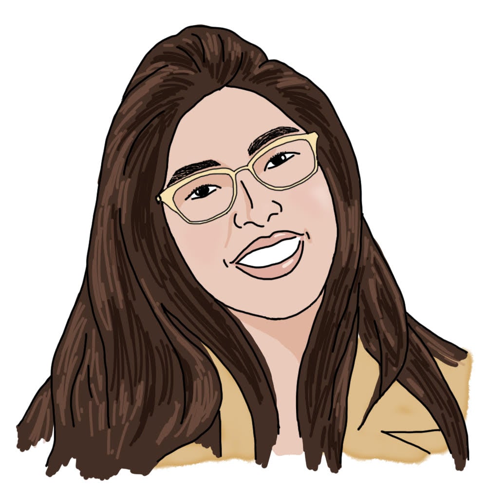 A head-and-shoulders illustration of Kano. She has medium brown skin, long brown hair and is wearing a darker yellow jacket. She has glasses and is smiling with her mouth open.