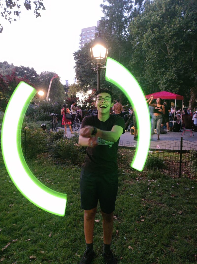 Me in a park. I am holding two glowstick poi which look like large circular swaths of light in the photo. I am grinning.