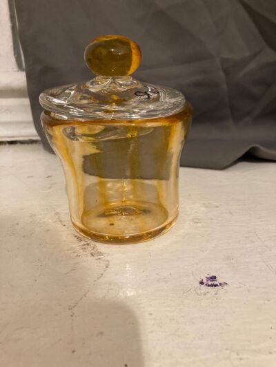 A jar with a cylindrical base that expands outwards at the rim. The jar is light yellow with darker yellow vertical stripes. It has a clear lid with a round handle that is the same yellow as the jar.
