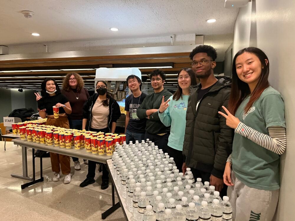 group of several mit students (lsc refreshment crew) smiling and stadning behind tables filled with popcorn and water bottles