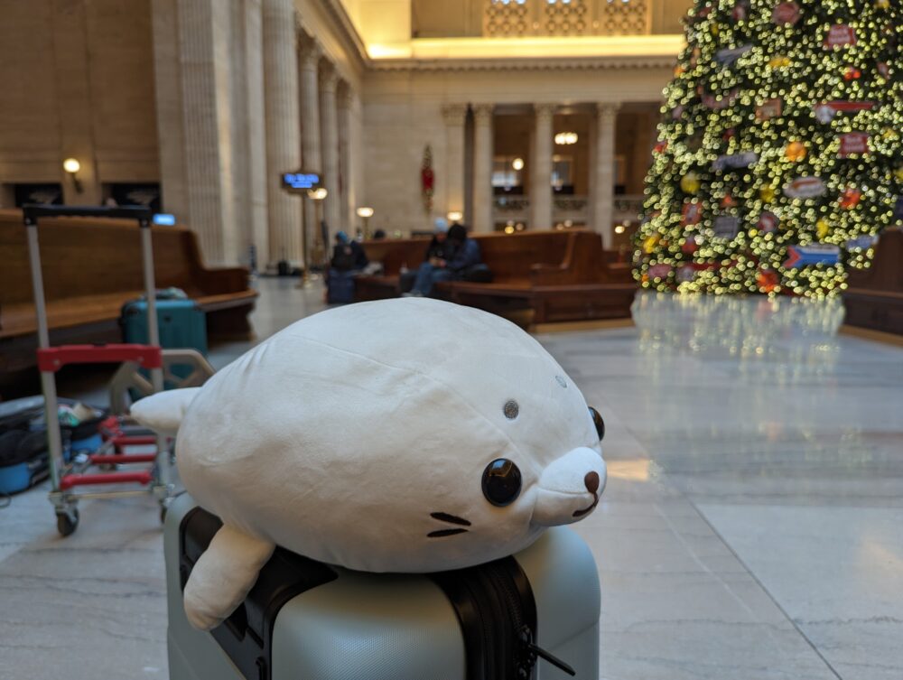 seal plushie on suitcase with train station in background, out of focus