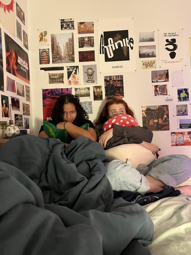 two people sitting on a bed in a dorm room hugging plushies