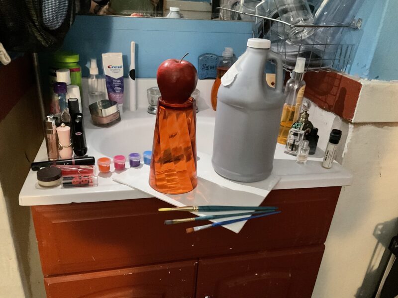 an apple on a cup on a canvas, next to a paint bottle and brushes