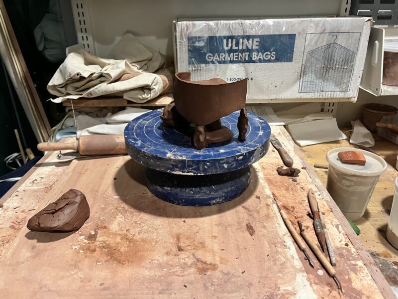 A picture of a ceramics workbench and lazy susan, on top of which is a brown half-formed clay vessel.