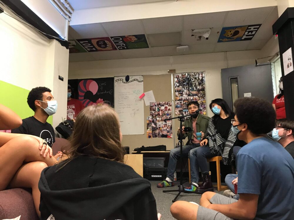 5-10 people gathered around an open mic night, where 2 people are performing
