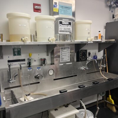 a picture of a long, deep, metal sink with various plastic tubs of chemicals on a shelf above it