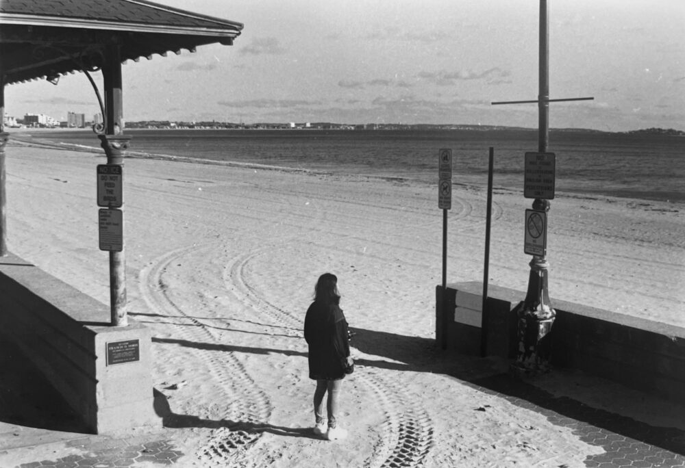 a black and white picture of a woman standing on sand, facing the ocean; she's sandwiched by two incomplete walls on either side