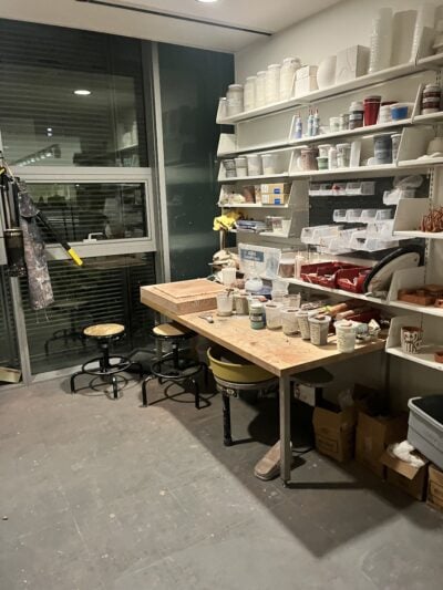 picture of a small ceramics studio, with a table in front of a wall of shelves. the shelves have various plastic cartons and boxes of chemicals, glazes, etc.