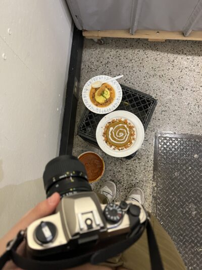 a point-of-view picture with a hand holding a film camera and two plates of food on a crate on the ground