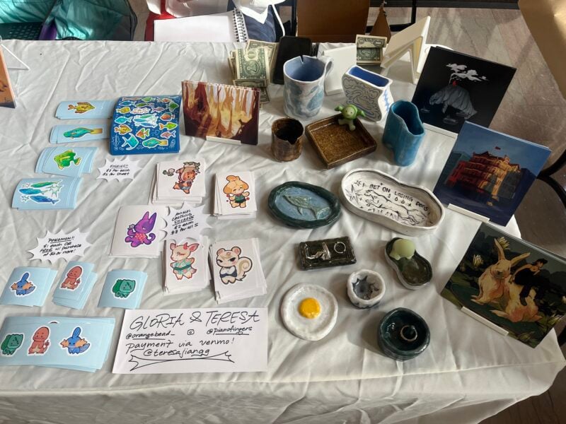 picture of a table with a white tablecloth covered in art prints, stickers, and ceramics