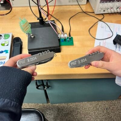 two hands, each holding silver utility knifes with "MIT 2.004" engraved on them