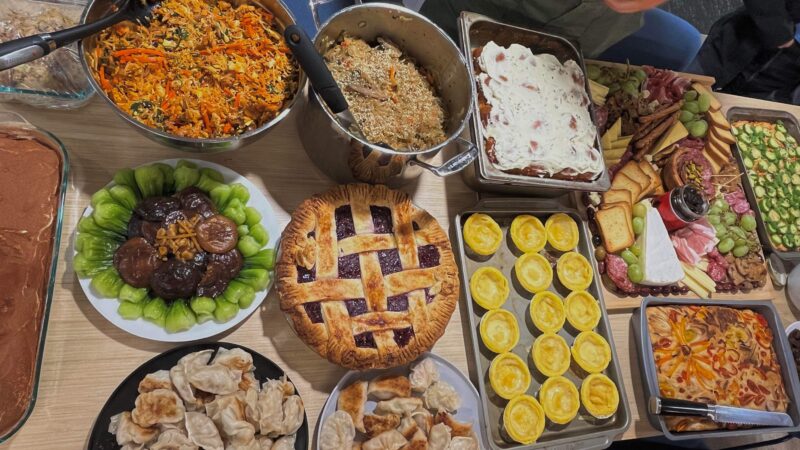 a picture of a table covered with plates of food. the food includes a charcuterie board, pie, pots of pasta and bakes, potstickers, egg tarts, tiramisu, and mushrooms.