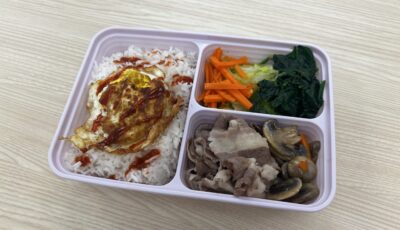 Image of bibimbap in meal prep container with three areas. First area has rice with egg on top and bibimbap sauce. Second compartment has sliced carrots and spinach/kale. Third has mushrooms and sliced beef.