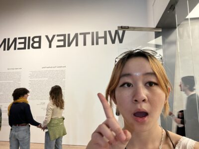 a girl taking a selfie points at text on the wall behind her. the text reads "whitney biennial" (partially cut off)