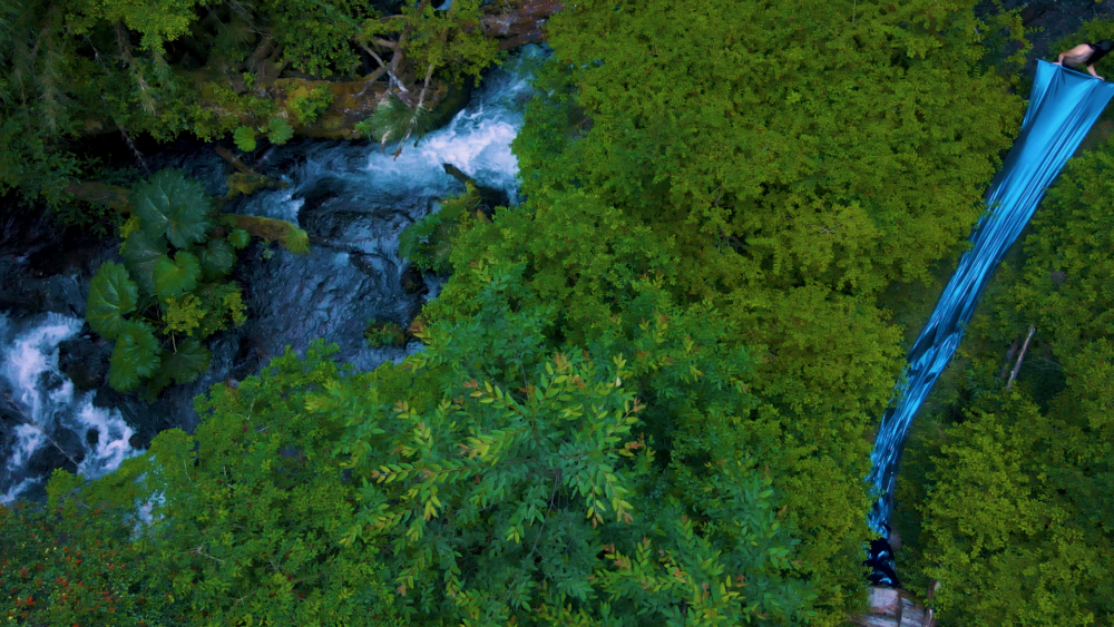 a still from seba's film. aerial view of a river winding through a forest, with a figure trailing metallic blue fabric through it.