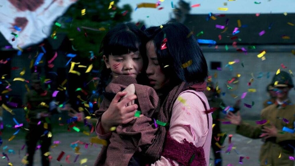 a still from a video. a woman hugs her baby, an expression of fear on her face, as confetti falls around her.