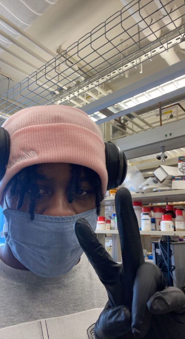 A selfie of Andre wearing a pink beanie, headphones, a mask, and a black rubber glove. He's standing in a lab and making a peace sign at the camera. 