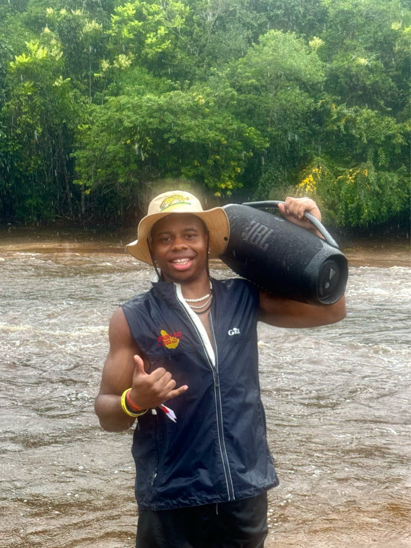 Andre standing in front of a river while it's raining, in a bucket hat and a vest, holding a big black speaker on one shoulder.