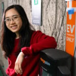 A young woman in glasses smiling, leaning against a wall near an EV charging station.