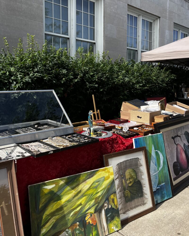 Image of red tablecloth table in front of bushes. Table has wooden boxes full of books, paintings, and jewelry.
