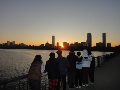 seven figures staring out at the sun rising over Boston skyline from the Harvard bridge