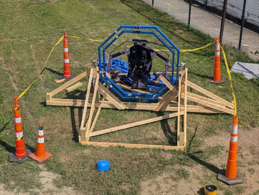 A wooden space trainer gyroscope ride surrounded by five orange cones and caution tape. 