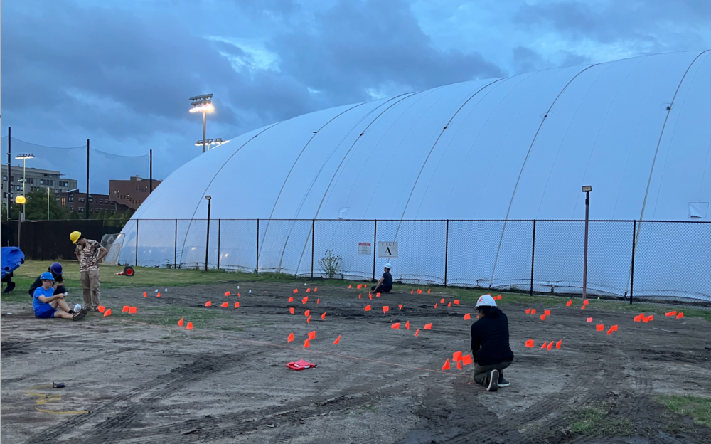 A person in a white hard hat, kneeling down in some dirt by orange flags that mark out the fort's footprint.