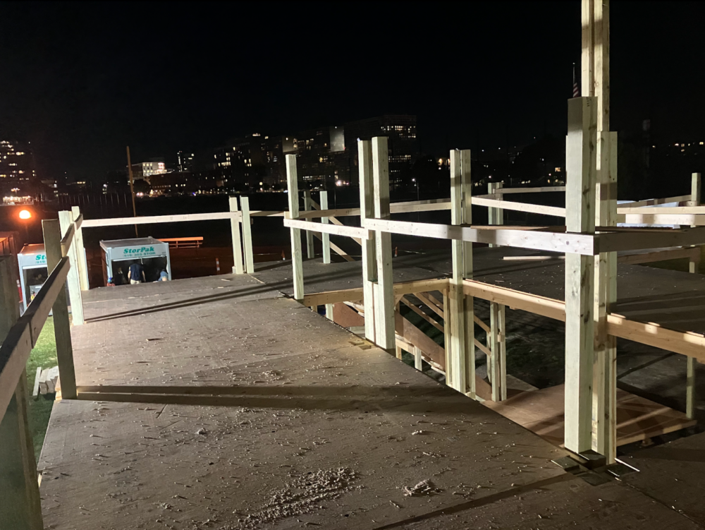 A picture of the second story of a wooden fort, with sawdust-sprinkled plywood, posts jutting upward, and the MIT campus in the background.