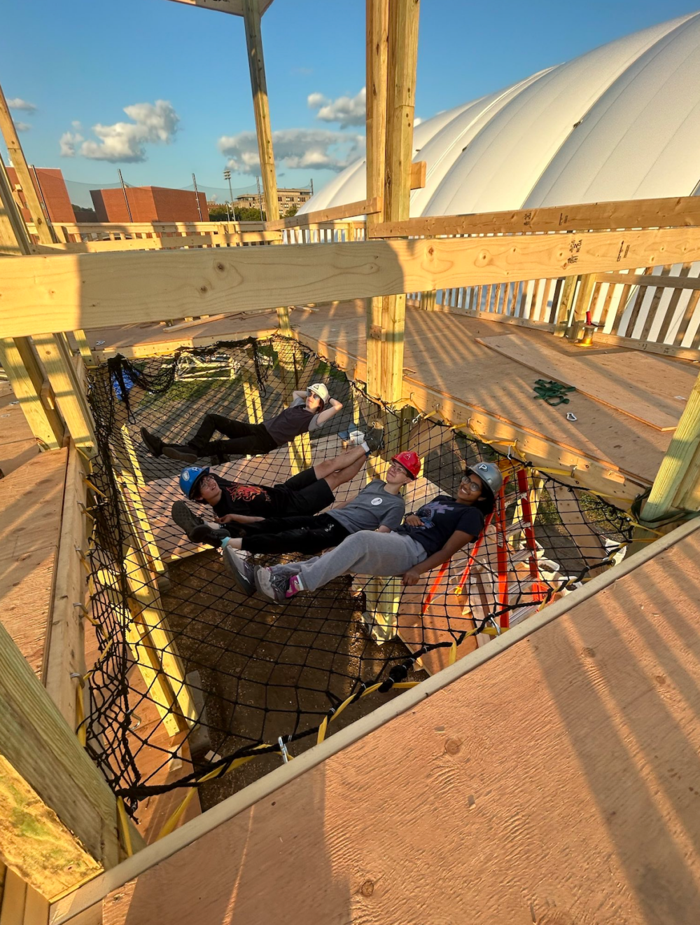 4 East Campus residents (and fort builders) in hard hats, laying in the fort's net.